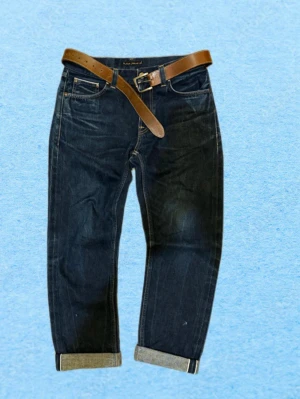 Mörkblå selvedge jeans från Nudie Jeans Co - Snygga mörkblå jeans från Nudie Jeans Co i rak modell med uppvikta benslut och klassiska fem fickor. Jeansen har kontrastsömmar och medföljer ett brunt läderbälte. Perfekta för en avslappnad och trendig look.