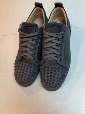 Gråa loubs - Snygga svarta sneakers med klassisk låg profil och röd sula. Skorna har en stilren design med svarta skosnören och diskreta detaljer. Perfekta för dig som gillar en clean look med en twist. Passar till både jeans och andra streetwear-outfits.
