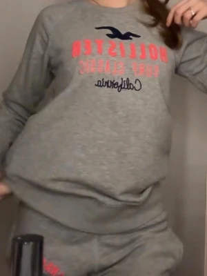 Grått Hollister tröja  - Mysig grå tröja från Hollister med tryck i rosa och mörkblått på både tröja och byxor. Setet har en avslappnad passform, rund halsringning och mjukt material som passar perfekt för chill dagar hemma.