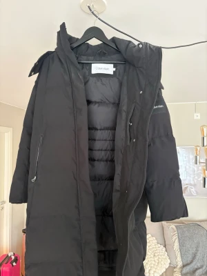 Svart lång dunjacka Calvin Klein - Säljer en svart lång dunjacka från Calvin Klein med huva och dragkedja. Jackan har diskret logga på ärmen och stora fickor med dragkedja. Perfekt för kalla vinterdagar. Riktigt bra skick använd max 10 gånger. 