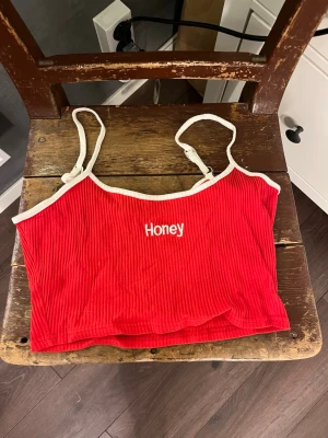Röd ribbad linnetopp med texten Honey - Trendig röd ribbad linnetopp med vita kanter och smala axelband. Framtill står det 'Honey' broderat i vitt. Perfekt för dig som vill ha en söt och enkel topp till sommaren.