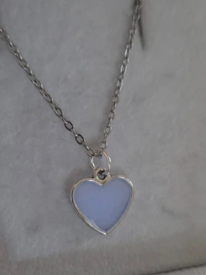 Halsband med självlysande hjärthänge - Snyggt halsband med ett hjärtformat hänge som lyser i blått i mörker och är vitt/creme/svagt glittrig i dagsljus. Kedjan är i rostfritt stål och hänget har en slät, glansig yta. Perfekt för dig som gillar unika accessoarer med cool effekt. Hjärtat började lysa i mörkare omgivning efter att jag tog kort på det i fönstret. (Vanligt dagsljus, även molnigt väder, aktiverar skenet i hänget).