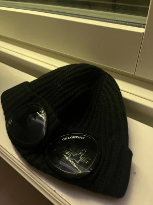 Svart C.P. Company mössa med goggles - Säljer en svart ribbstickad mössa från C.P. Company med goggles framtill. Mössan har en bred uppvikt kant och är tillverkad i mjukt stickat material. Passar för en tonåring. Mössan är i nyskick och använd fåtal gånger. 