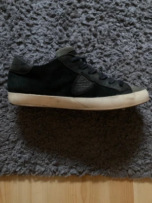 Svarta Phillip Model - Snygga svarta sneakers i mocka med vita sulor och svarta skosnören. Klassisk låg modell med diskret detalj på sidan och mjuk plös. Passar perfekt till jeans eller streetwear.Två små skador på insidan av skorna 
