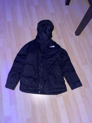 Svart vindjacka från The North Face - Snygg svart vindjacka från The North Face med huva och dragkedja framtill. Jackan har två sidofickor och logga på bröstet. Tillverkad i slitstarkt material som står emot vind och lätt regn. Perfekt för dig som vill ha en stilren och funktionell jacka.