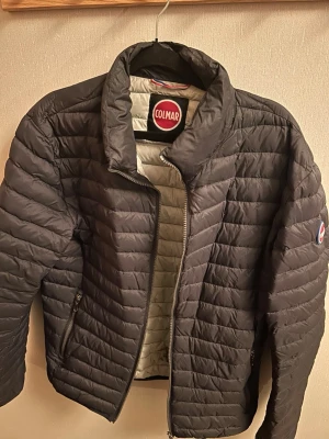 Svart dunjacka från Colmar - Snygg svart dunjacka från Colmar med quiltad design och hög krage. Jackan har dragkedja framtill, två sidofickor med dragkedja och Colmar-logga på ärmen. Insidan är ljusgrå och materialet är lätt och smidigt, perfekt för kyliga dagar.