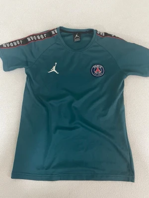 PSG x Jordan blå träningströja - Snygg blå träningströja från PSG x Jordan med rund hals och korta ärmar. Tröjan har vita Jordan-loggan på bröstet och PSG-emblemet. Axelbanden har svart och röd detalj med texten 'JORDAN'. Perfekt för fotbollsträning eller gymmet.