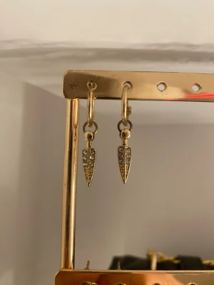 Guldiga örhängen med glittriga stenar - Säljer ett par snygga guldiga örhängen med små glittriga stenar på spetsiga hängen. Perfekta för dig som vill ha något stilrent men ändå med lite bling. Passar till många olika looks och är lätta att matcha.