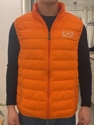 Orange dunväst från Emporio Armani EA7 - Snygg orange dunväst från Emporio Armani EA7 med quiltad design och dragkedja framtill. Västen har hög krage och två sidofickor. EA7-logga i vitt på bröstet ger en sportig vibe. Perfekt att slänga över en hoodie eller tröja för extra värme. Västen är egentligen i storlek XL men den sitter som S-M. Jag är 170 och den är för lång för mig.