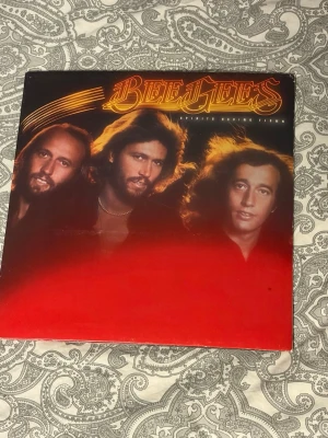 Bee Gees vinylskiva - Vinylskiva med Bee Gees, ikoniskt omslag i rött och svart med gruppens logga i guld. Inuti finns ett uppvikbart konvolut med bilder på bandmedlemmarna och låttexter tryckta i rött. Perfekt för samlare av klassisk pop och disco.