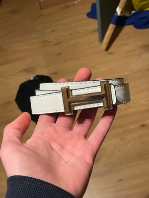 Vitt  bälte med guldfärgat H-spänne - Snyggt vitt bälte med det ikoniska H-spännet i guldfärg. Bältet är gjort i skinn och har en stilren, klassisk design som passar till många olika outfits. Perfekt accessoar för att lyfta din look.