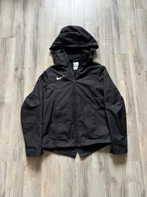 Svart vindjacka från Nike - Snygg svart vindjacka från Nike med hel dragkedja framtill och huva. Jackan har en minimalistisk design med diskret Nike-logga på bröstet och är tillverkad i lätt, vindtätt material. Perfekt för blåsiga dagar och enkel att matcha med olika outfits. Den är stilig och passar även 160-165 cm.