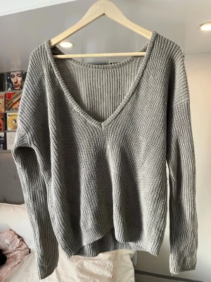 Grå stickad tröja med v-ringning - Snygg grå stickad tröja med djup v-ringning framtill och rundad halsringning bak. Tröjan har långa ärmar och är ribbstickad i ett mjukt material, perfekt för lager på lager. Passar till både jeans och kjol för en avslappnad look.