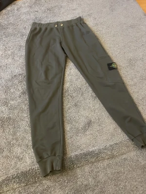 Gröna mjukisbyxor från Stone Island - Snygga gröna mjukisbyxor från Stone Island med ikonisk patch på benet. Byxorna har resår i midjan med två knappar, muddar vid bensluten och en ficka på sidan. Perfekta för en chill och avslappnad stil.