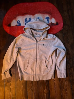 Grå hoodie från Polo Ralph Lauren - Snygg grå hoodie från Polo Ralph Lauren med dragkedja framtill och två fickor. Klassisk broderad logga på bröstet och justerbar huva. 