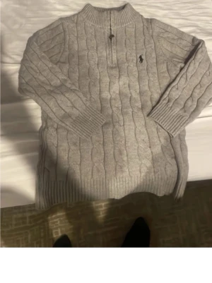 Grå kabelstickad tröja Polo Ralph Lauren - Kabelstickad grå tröja från Polo Ralph Lauren med halv dragkedja och ribbade muddar. Klassisk logga broderad på bröstet. Tröjan har hög krage och lång ärm, perfekt för kyliga dagar. Materialet känns mjukt och håller värmen.