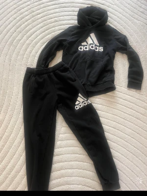 Svarta mjukisbyxor från Adidas - Säljer ett par svarta mjukisbyxor från Adidas med vit logga på vänster lår. Byxorna har resår i midjan och muddar vid bensluten. Tillverkade i mjukt och bekvämt material, perfekta för chill dagar. Klassisk sportig look med fickor på sidorna.