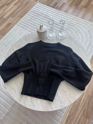 Svart figursydd tröja - Trendig svart stickad tröja med figursydd passform och dekorativa knappar framtill. Tröjan har vida ärmar och ribbade muddar vid halsen. Perfekt för lager på lager och enkel att styla till olika outfits.