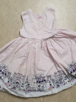 Klänning - Söt ljusrosa omlottklänning med vid kjol och ett unikt stadsmotiv i lila, svart och vitt längs fållen. Klänningen är ärmlös och har en omlottdesign upptill. Perfekt för dig som gillar lekfulla detaljer och mjuka pasteller. Har gul fläck.