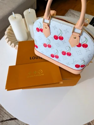 Iouis Vuitton handväska  - Supersöt vit handväsk med körsbärsmönster och beige detaljer. Väskan har en stor rosett framtill, guldfärgade metalldetaljer och röd insida. Perfekt storlek för det viktigaste och har både handtag och axelrem  .                                                                            .
