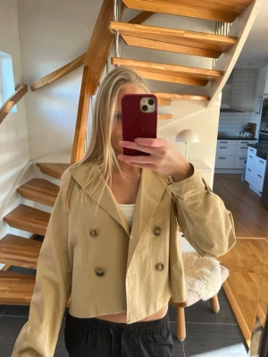 Kort beige trenchcoat från H&M - Stilren kort trenchcoat i beige från H&M med dubbelknäppning och bred krage. Jackan har långa ärmar och en rak, boxig passform som ger en modern look. Tillverkad i ett lätt tyg som passar perfekt till vår och höst.