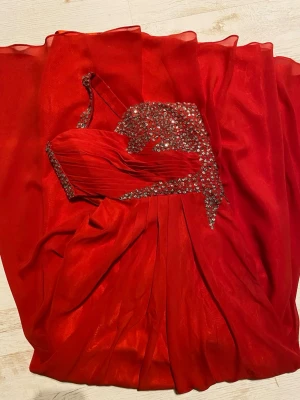 Röd oneshoulder långklänning med paljetter - Använd en gång. Lång balklänning med paljetter på överdelen. Klänningen har en slits på ena sidan av kjolen. Överdelen är draperad och har en asymmetrisk axelbandsdesign. Klänningen har en dragkedja i sidan. Längd 138cm, Bröst 84 cm, midja 77 -80 cm. 