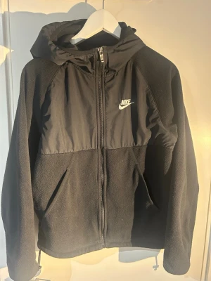 Svart fleecejacka med huva från Nike - Säljer en svart fleecejacka från Nike med huva och dragkedja framtill. Jackan har två fickor, broderad Nike-logga på bröstet och är tillverkad i mjuk fleece. Perfekt för kyliga dagar och enkel att matcha med allt.
