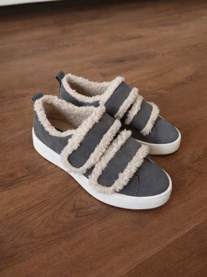 Grå sneakers med fuskpälsdetaljer - Unika grå/bruna sneakers ifrån Duffy med två breda kardborreband och fluffiga fuskpälsdetaljer längs kanterna och insidan. Skorna har en vit sula och är tillverkade i mockaimitation. Perfekta för dig som vill ha något extra och mysigt till din stil. Använd max 2 ggr! Nypris 900