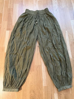 Olivgröna crinkle mjukisbyxor - Säljer ett par olivgröna baloongbyxor från Zimmermann  med crinkle-effekt och resår i midjan samt benslut. Byxorna har dragsko med träpärlor i midjan för extra detalj. Luftig och avslappnad passform. 