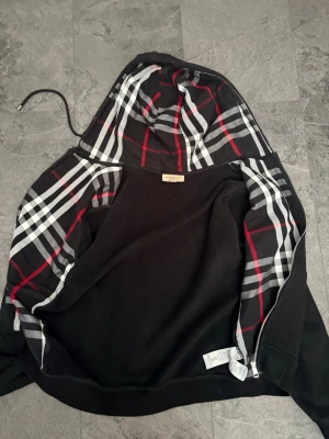 Svart hoodie från Burberry med rutigt foder - Snygg svart hoodie från Burberry med det klassiska rutiga fodret i huvan och på insidan. Dragkedja framtill och mjukt material som känns skönt mot huden. Perfekt för dig som vill ha en stilren hoodie med exklusiv känsla och ikoniskt mönster.