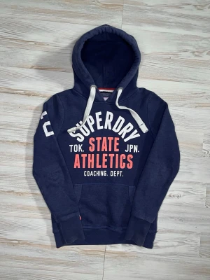 Mörkblå hoodie från Superdry - Mörkblå hoodie från Superdry med vit och röd text på bröstet och siffran 2 på ärmen🤩 hoodien passar nån i Xs-S och är i fint skick🙌🏻 Hör av er vid funderingar samt snabbaffär😇