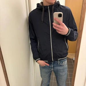 Mörkblå vindjacka med huva - Snygg mörkblå vindjacka med huva och dragsko. Jackan har vita detaljer längs dragkedjor och muddar, samt två fickor med dragkedja. Tillverkad i lätt och tunt material, perfekt för blåsiga dagar. Passar till jeans och streetwear.