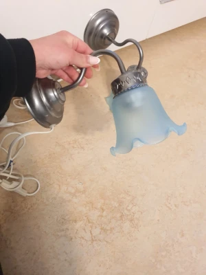 Vägglampor med blått glas - Två snygga vägglampor i metall med dekorativa, vågiga lampskärmar i frostat blått glas. Lamporna har rund väggplatta och böjd arm i silverfärgad metall. Perfekt för att ge rummet en retro eller vintagekänsla.