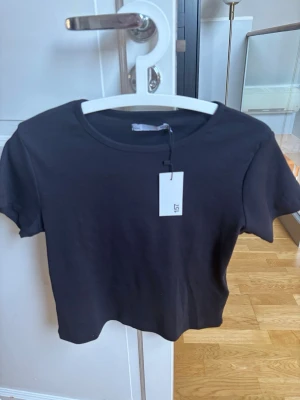 Svart croppad t-shirt från 157 - En enkel och stilren svart croppad t-shirt från 157. T-shirten har rund halsringning och korta ärmar. Tillverkad i mjuk bomull som känns skön mot huden. Perfekt till jeans eller kjol för en clean look.