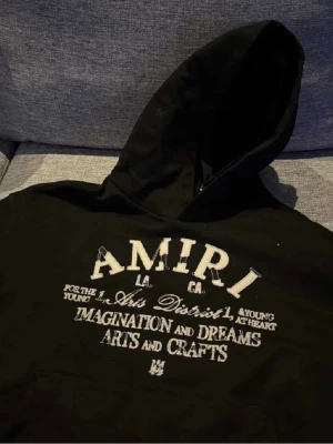 Svart Amiri hoodie    - Säljer en svart hoodie från Amiri med stor vit broderad text och tryck på bröstet. Hoodien har klassisk huva och dragsko samt detaljerade sömmar. Materialet är mjukt och bekvämt, perfekt för en avslappnad streetwear-look.