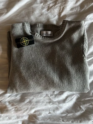 Grå stickad tröja från Stone Island - Snygg grå stickad tröja från Stone Island med klassisk rund hals och ribbade muddar. Storlek junior 12.  Tröjan har den ikoniska svarta logotypen med kompass på ärmen. Perfekt för dig som gillar stilrena och tidlösa plagg. Tveka inte att höra ev er vid frågor🙌