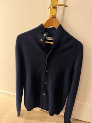 Mörkblå kashmirkofta från Soft Goat - Snygg mörkblå kofta från Soft Goat i mjuk kashmir. Rak passform med hög krage och knappar hela vägen framtill. Perfekt till lager på lager och riktigt skön mot huden. Tröjan är i väldigt bra skick!