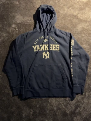 Mörkblå Yankees hoodie från Nike - Snygg mörkblå hoodie från Nike med New York Yankees-tryck på bröstet och logga på ärmen. Klassisk känguruficka framtill och justerbar huva med snören. Perfekt för dig som gillar sportig streetwear och vill representera Yankees.