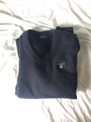 Svart stickad tröja från Gant - Klassisk svart stickad tröja från Gant med V-ringning och broderad logga på bröstet. Tröjan har lång ärm och är tillverkad i mjukt material som passar perfekt till lager på lager. Enkel och stilren design som funkar till det mesta.