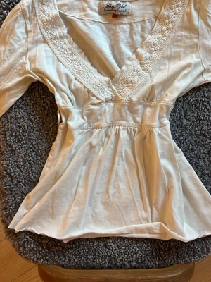 Vit babydoll top - Söt cremefärgad babydoll top från Stars She med v-ringning och spetsdetaljer längs ringningen. Finns två små snören vid midjan så den var tidigare knytbar (köpt second hand), och den är nedklippt längst ner så det kan vara lite grova kanter. 