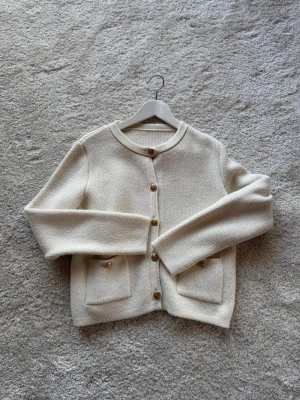 Krämvit kofta med guldknappar - Stilren krämvit kofta i stickat material med rund halsringning och dekorativa guldknappar framtill. Två praktiska fickor framtill ger extra detaljer. Perfekt för dig som gillar en clean och klassisk look. Sällan använd. 