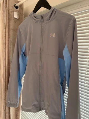 Gråblå träningsjacka Under Armour - Snygg träningsjacka från Under Armour i grått med ljusblå detaljer längs sidorna och ärmarna. Jackan har huva, hel dragkedja framtill och elastiska muddar vid ärmsluten. Perfekt för löpning eller gym. Tillverkad i lätt funktionsmaterial. Den säljs inte längre har haft på mig den några gånger 