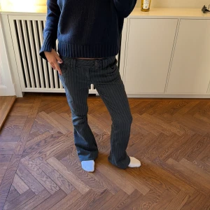 Blåa pinstripe jeans med låg midja - Säljer ett par gråa jeans med vita pinstripes och låg midja. Jeansen har raka ben och klassisk femficksmodell. Snygg detalj med bälteshällor och knappgylf. Perfekta för en trendig och avslappnad look.