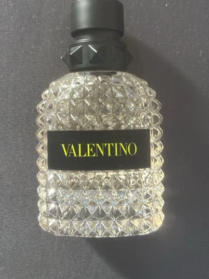 Valentino parfym med nitar och glas - Exklusiv parfym från Valentino i en transparent, diamantmönstrad glasflaska med svart etikett och gul text. Korken är svart med dekorativa nitar som ger en lyxig och modern känsla. Perfekt för dig som gillar eleganta och trendiga dofter. Mer än hälften finns kvar! 