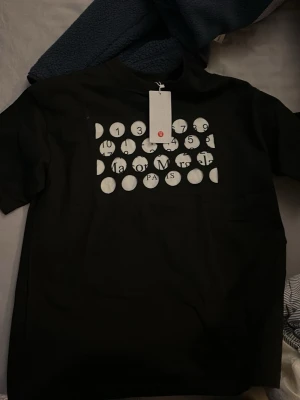 Svart t-shirt från Maison Margiela - Cool svart t-shirt från Maison Margiela med vit grafisk print på bröstet som består av cirklar med siffror och text. Klassisk passform och korta ärmar, perfekt för dig som gillar streetwear och designerplagg.