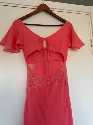 Lång rosa klänning med spetsdetaljer - Snygg långklänning i rosa med volang-ärmar. Klänningen har spetsdetaljer i midjan, öppen rygg och snörning i ryggen 