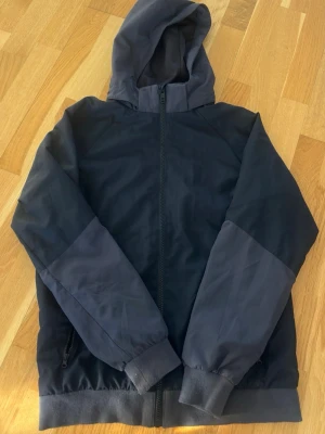 Svart dunjacka med huva - Svart dunjacka med huva och hel dragkedja framtill. Jackan har ribbade muddar vid ärmslut och nederkant samt två sidofickor med dragkedja. Enkel och stilren design som håller dig varm under kalla dagar.