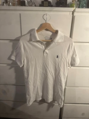 Vit pikétröja från Polo Ralph Lauren - Klassisk vit pikétröja från Polo Ralph Lauren med blå broderad logga på bröstet. Tröjan har krage, korta ärmar och knäppning framtill. Modellen är slim fit och tillverkad i bomull för en skön känsla.