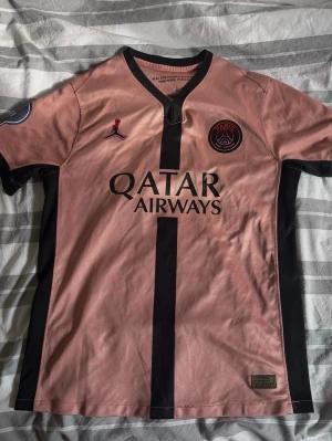 PSG rosa matchtröja Dembélé #10 - Säljer en rosa PSG matchtröja med svarta detaljer, Jordan-logga och Qatar Airways-tryck. På ryggen står O. Dembélé och nummer 10. Tröjan har korta ärmar och är tillverkad i ett lätt, ventilerande material perfekt för fotboll.