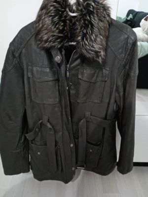 Svart skinnjacka med fuskpäls - Snygg svart skinnjacka med stor krage i fuskpäls och flera praktiska fickor framtill. Jackan har bälte i midjan och detaljerade sömmar på axlarna. Perfekt för dig som vill ha en cool och trendig look.